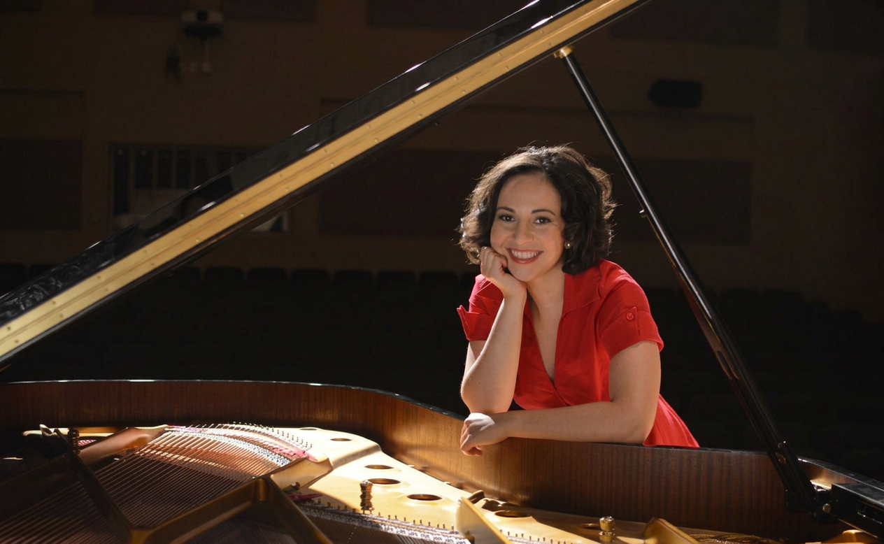 Dr. Monique Arar in a red dress, leaning on the open lid of a concert grand piano